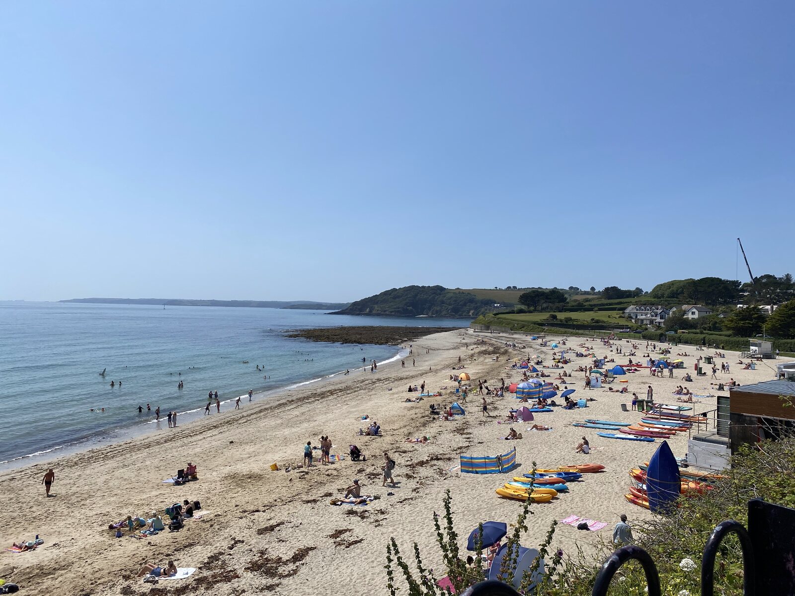 Gyllyngvase Beach in Falmouth on a busy summer’s day, a curved sandy bay with sunbathers and swimmers, and stacks of colourful kayaks and paddleboards ready for hire on the right.