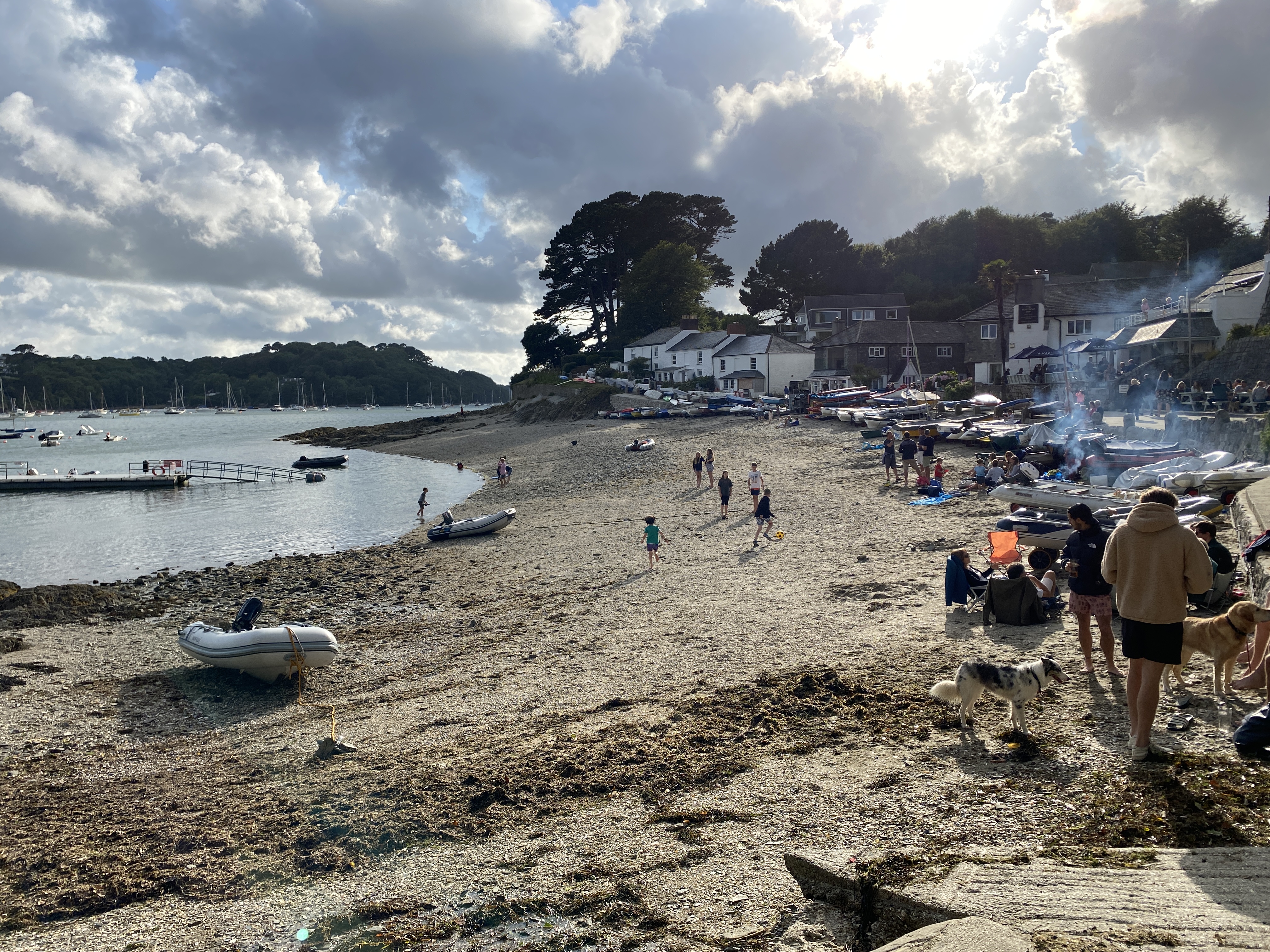 Boats pulled up on Helford Passage beach with people drinking and chatting outside the Ferry Boat Inn behind.