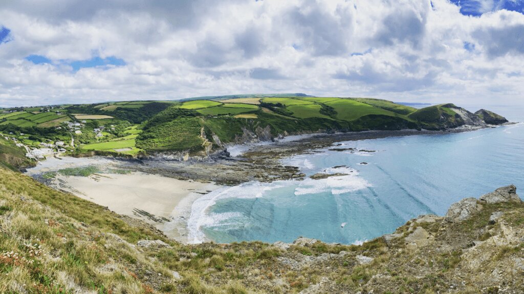 A stunning view of Crackington Haven, with towering slate cliffs, a pebble-strewn beach, and Atlantic waves crashing against the rugged North Cornwall coastline.