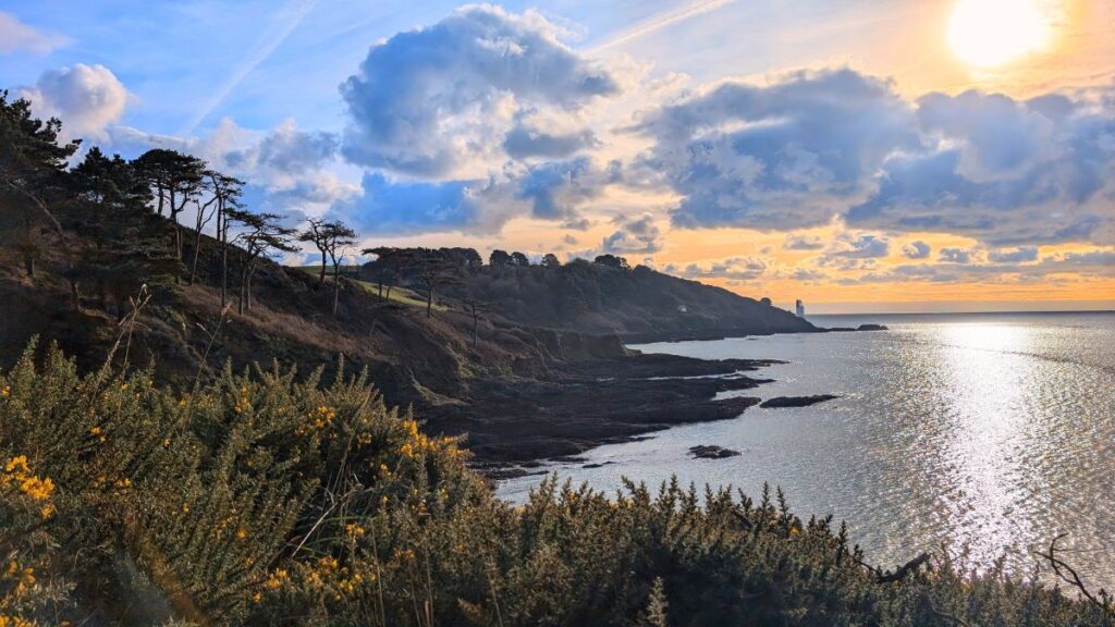 A coastal view looking towards St Anthony's Head and its lighthouse on the Roseland Peninsula, with rugged cliffs and blue waters stretching into the distance.