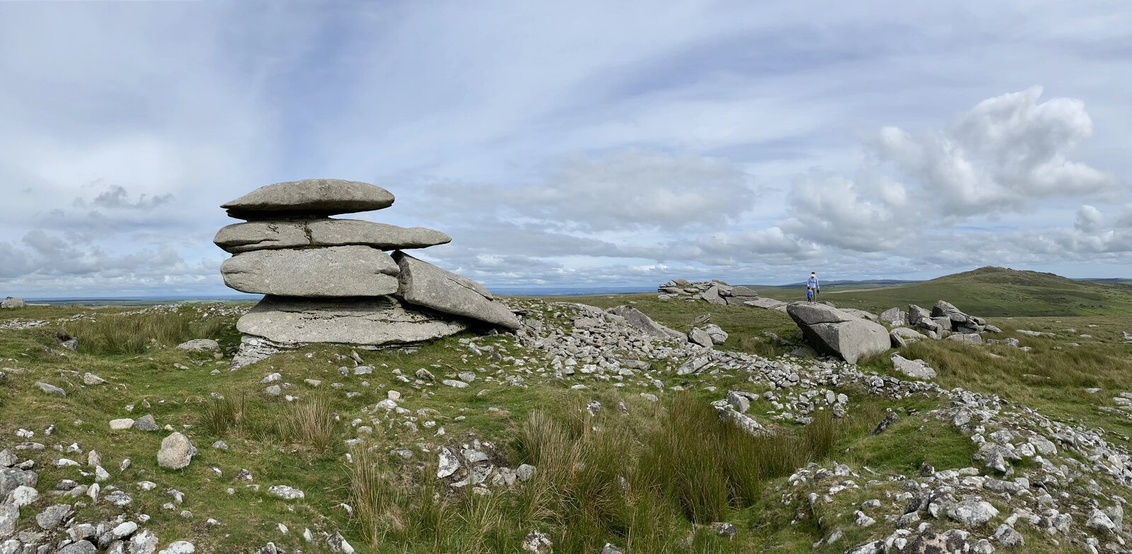 A stacked granite tor of balanced slabs on the Rough Tor ridge, likely Showery Tor, with rough grass and scattered boulders around the base and a second tor visible on the moor in the distance, two figures walking past for scale.