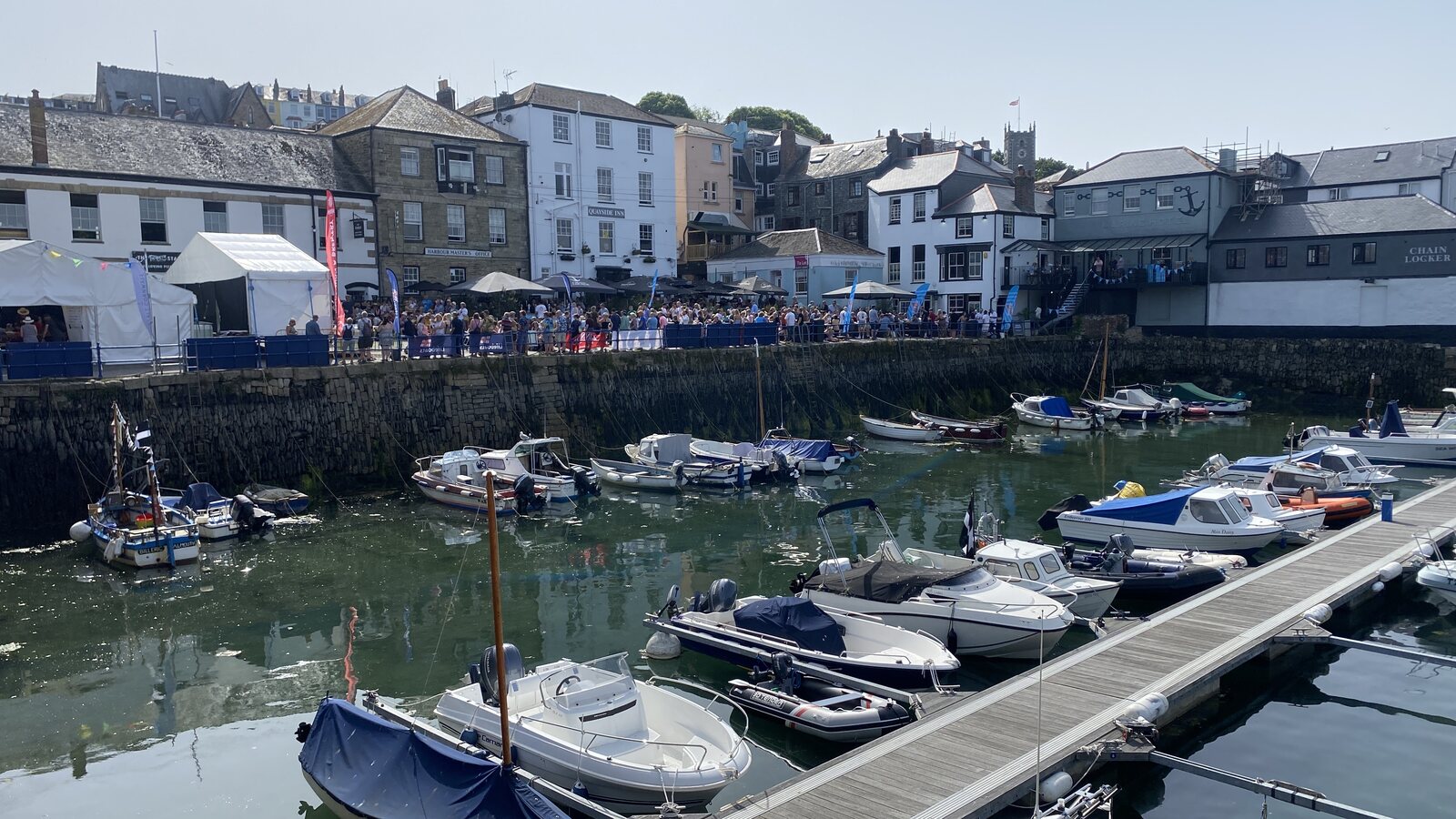 Custom House Quay in central Falmouth on a busy summer afternoon during what looks like a regatta day: a packed crowd of people on the harbour-front in front of the Quayside Inn, the Chain Locker pub and the old harbour-master's office, with small boats and dinghies moored in the inner harbour in the foreground.