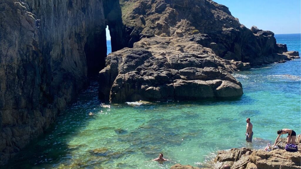 Nanjizal Beach near Land's End, with the Song of the Sea rock arch and a clear turquoise pool below where two people are swimming.