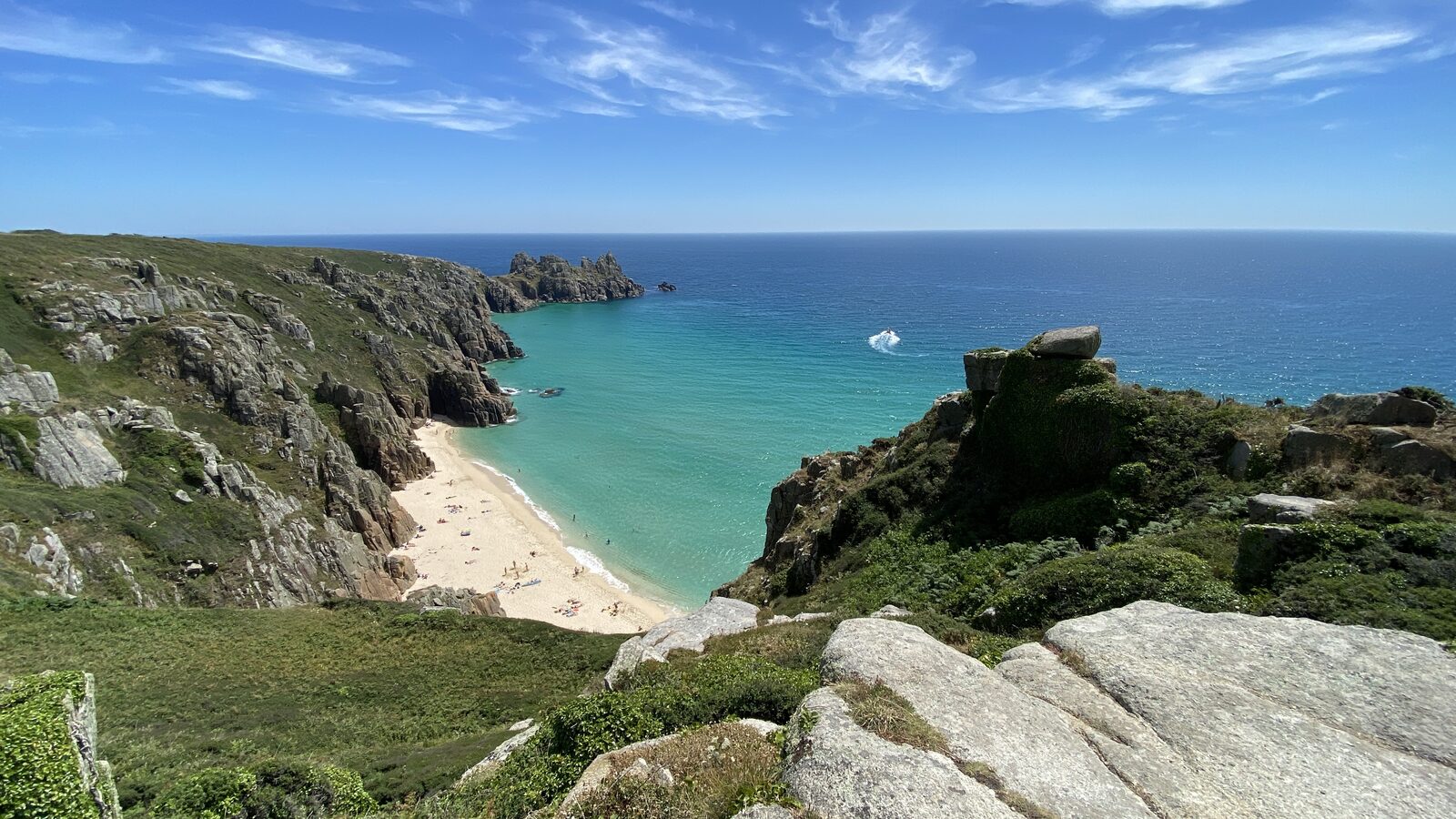 Pedn Vounder beach seen from the cliff path above, with white sand reaching into turquoise water and Logan Rock on the headland in the distance.