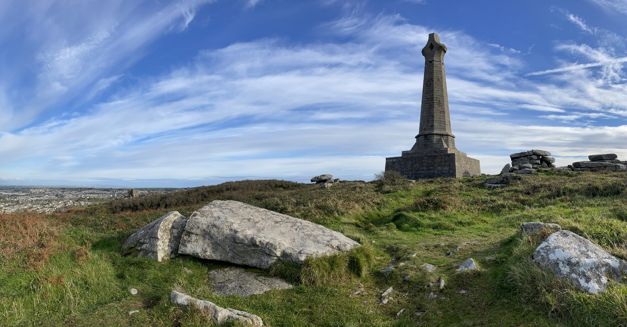 The Basset Monument, a tall granite obelisk topped with a Celtic cross, on the summit of Carn Brea above Redruth, with Carn Brea Castle visible in the distance to the left.