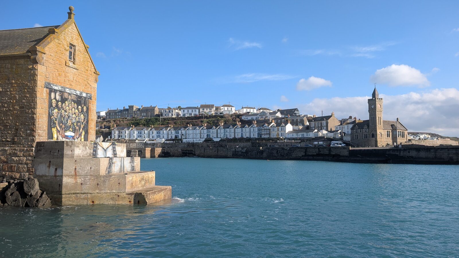 Porthleven harbour on a sunny day, with the historic Bickford-Smith Institute clock tower on the right and the lifeboat house mural on the left, calm turquoise water in the harbour and the row of seafront houses behind.