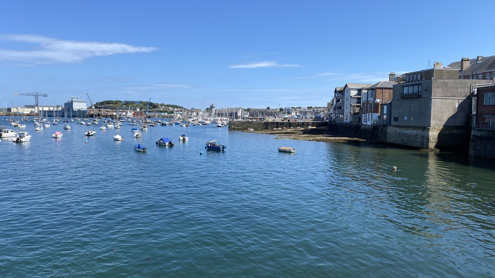 The view looking back across Falmouth's inner harbour from the pier at the central town end (near The Moor and Chintz), towards Pendennis Headland in the distance: small motor launches and dinghies moored in the calm foreground water, the cranes of Falmouth Docks visible in the middle distance, and the harbour-front town buildings rising on the right.