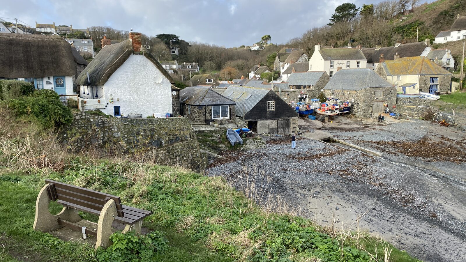Cadgwith fishing village in winter, with thatched cottages, granite buildings, the working fishing fleet pulled up on the slipway, and a wooden bench in the foreground.