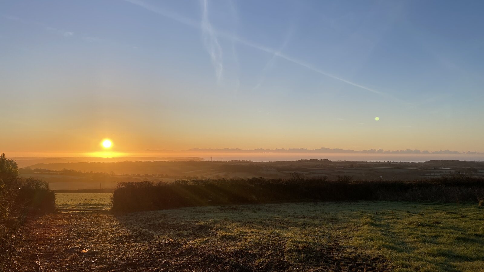Sunrise from the high ground above Mabe parish, looking south-east across rolling fields and Cornish hedges towards Falmouth Bay in the distance: the rising sun low and golden in the sky, throwing layered light across a landscape of hedge-lined fields and distant headlands, with the calm sea visible on the far horizon.