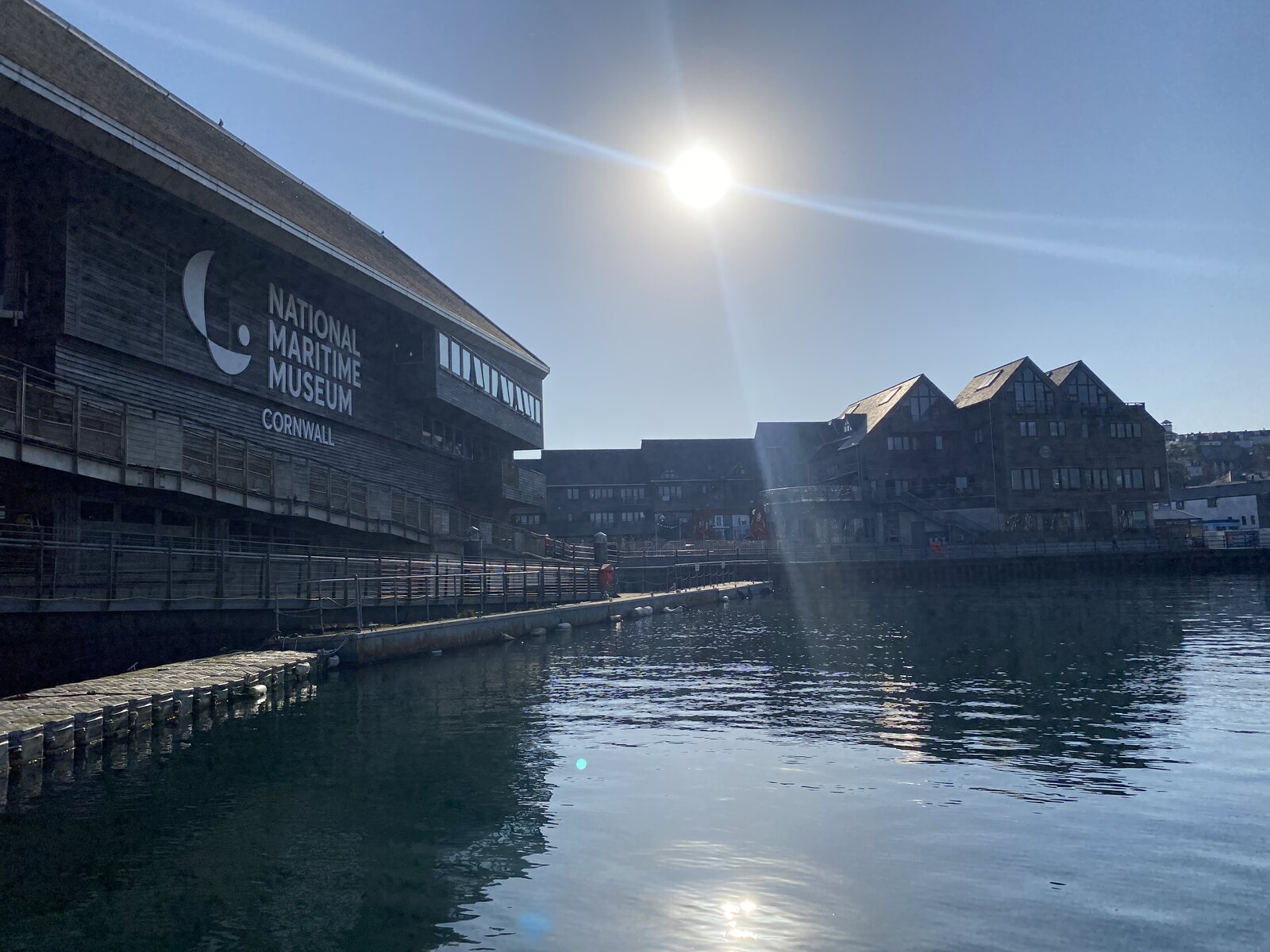 The exterior of the National Maritime Museum Cornwall on Discovery Quay in Falmouth, with the timber-clad building rising over the harbour and the sun catching the water in the foreground.
