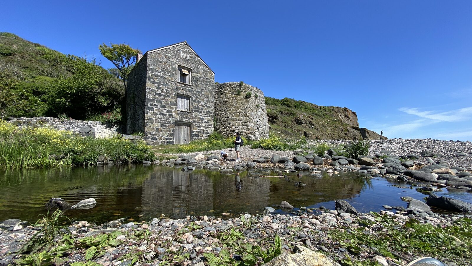 The 19th-century serpentine factory ruins at Carleon Cove on the Lizard, a stone industrial building reflected in a calm tidal pool with the cove and cliffs behind on a sunny summer day.