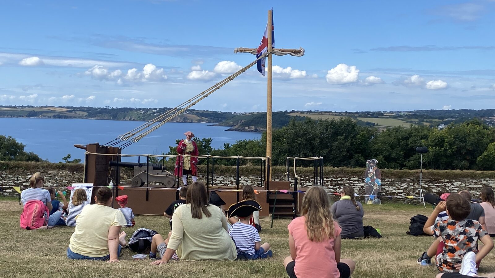 A pirates-themed family event at Pendennis Castle, with children and adults sitting on the grass watching a costumed performer on a wooden ship-deck stage with a Royal Navy flag, Falmouth Bay and the Roseland Peninsula visible in the background.
