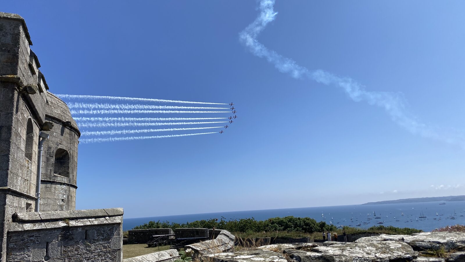 The Red Arrows displaying over Falmouth Bay seen from Pendennis Castle, with the Tudor stone keep on the left, smoke trails arcing across the sky, and yachts anchored in the bay below.