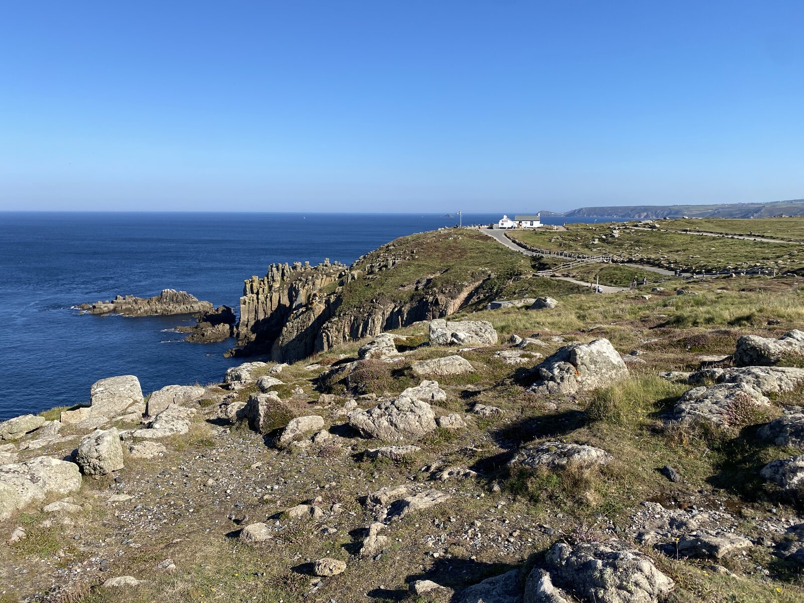 The dramatic granite cliffs at Land's End on Cornwall's far western tip, with the Atlantic sea breaking against the rocks below, the headland reaching out into the open sea and the Longships Lighthouse visible offshore in the distance.