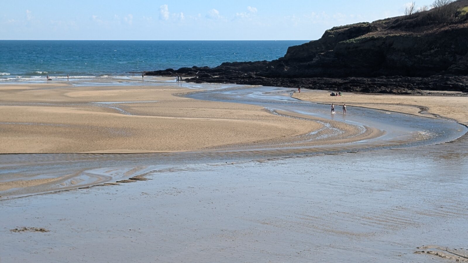 Maenporth Beach near Falmouth on a clear spring morning at low tide: wide expanses of clean ribbed sand with shallow tidal channels running across the foreground, a curving headland with rocky outcrops on the right, and a deep blue sea under a calm blue sky with a few people walking on the wet sand in the middle distance.