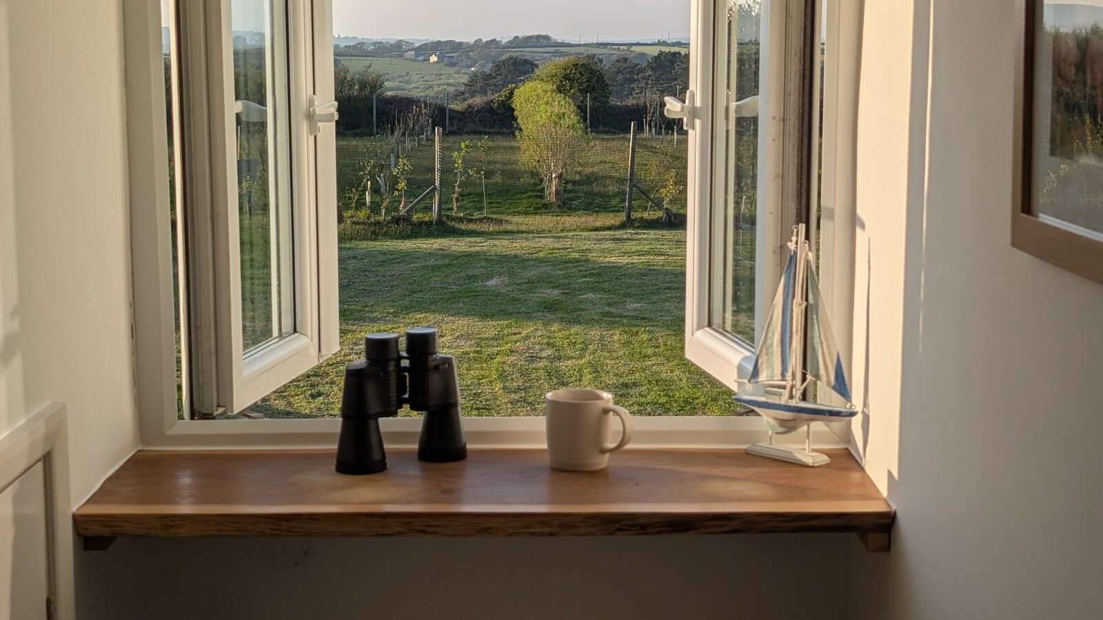 Looking out from the kitchen window of the Pigsty cottage at Trewena on an early-spring evening: an open sash window with a pair of binoculars and a mug of tea on the polished wooden sill, a small wooden model sailing boat to the right, and the view across the green orchard and meadow to the rolling Cornish countryside in the distance under a soft evening light.