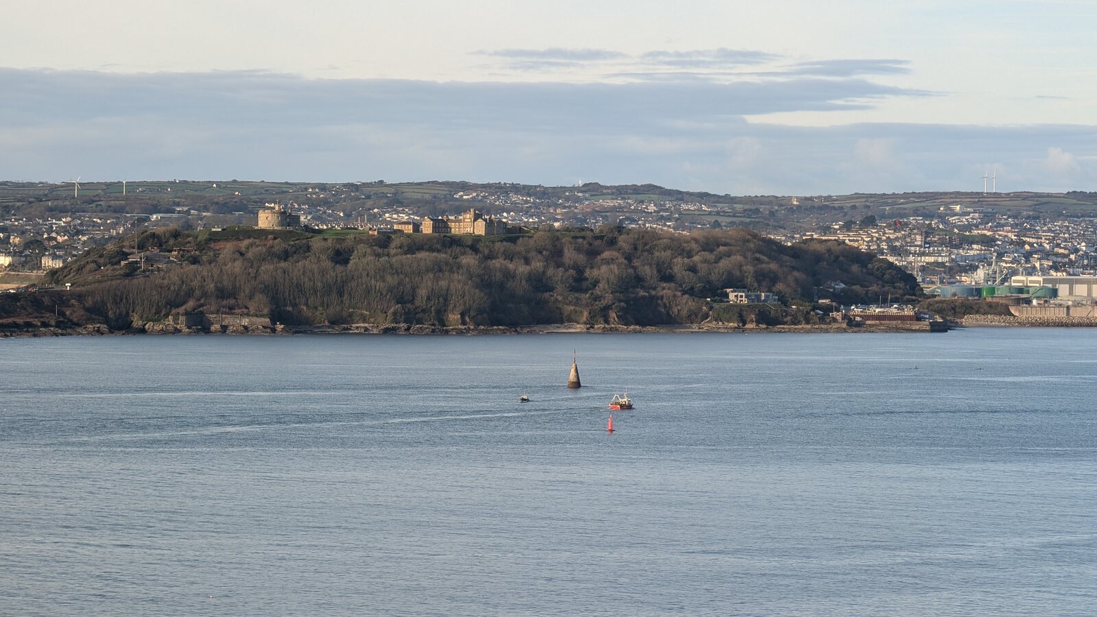 Pendennis Castle on its wooded headland across the calm water of the Fal, with Falmouth behind and a small red fishing boat passing a channel marker.