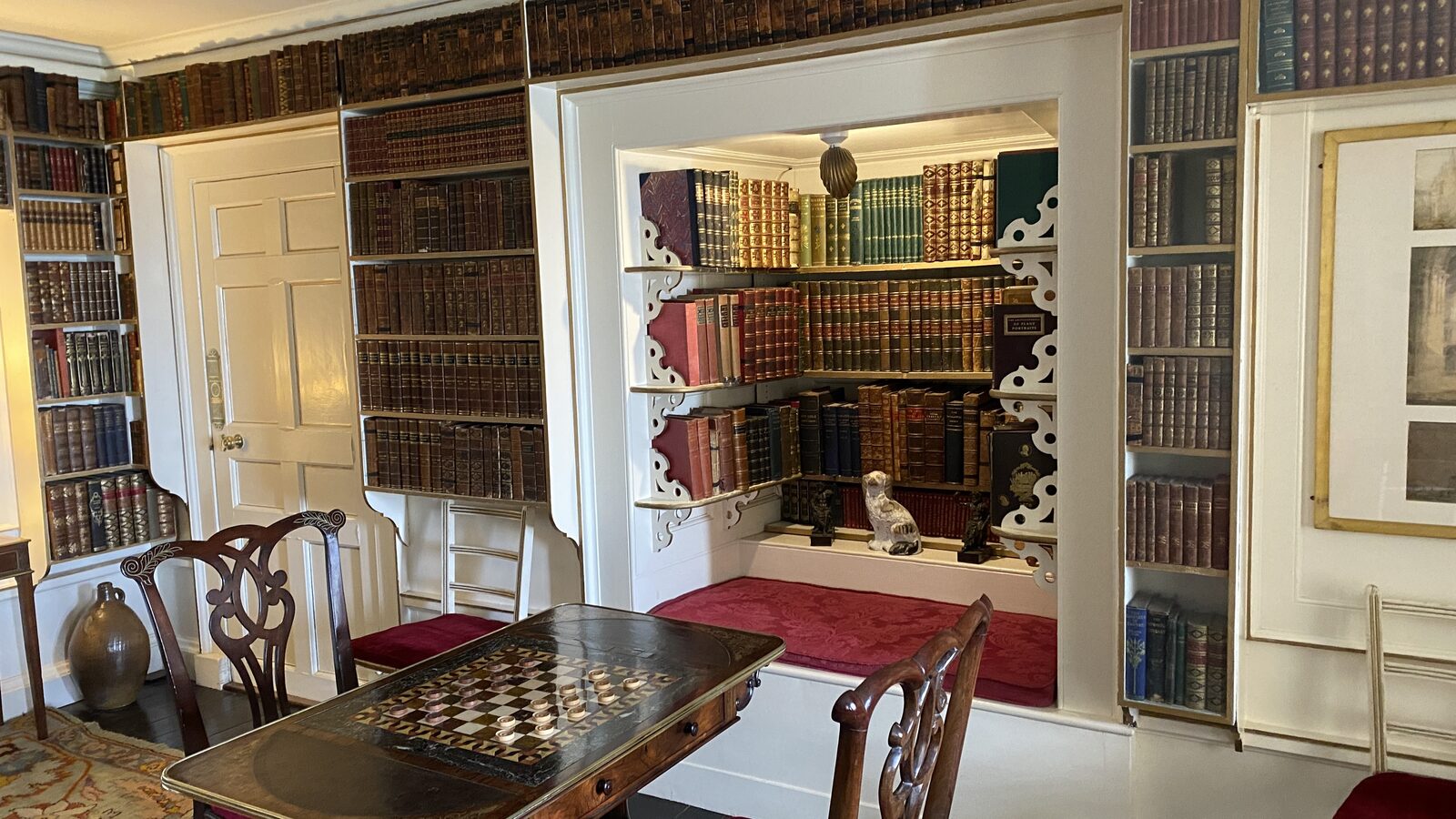 The library at St Michael's Mount castle, with floor-to-ceiling antique bookshelves of leather-bound volumes, a games table set with a draughts board mid-game, Chippendale chairs, and a red velvet window-seat alcove framed by ornate white fretwork.