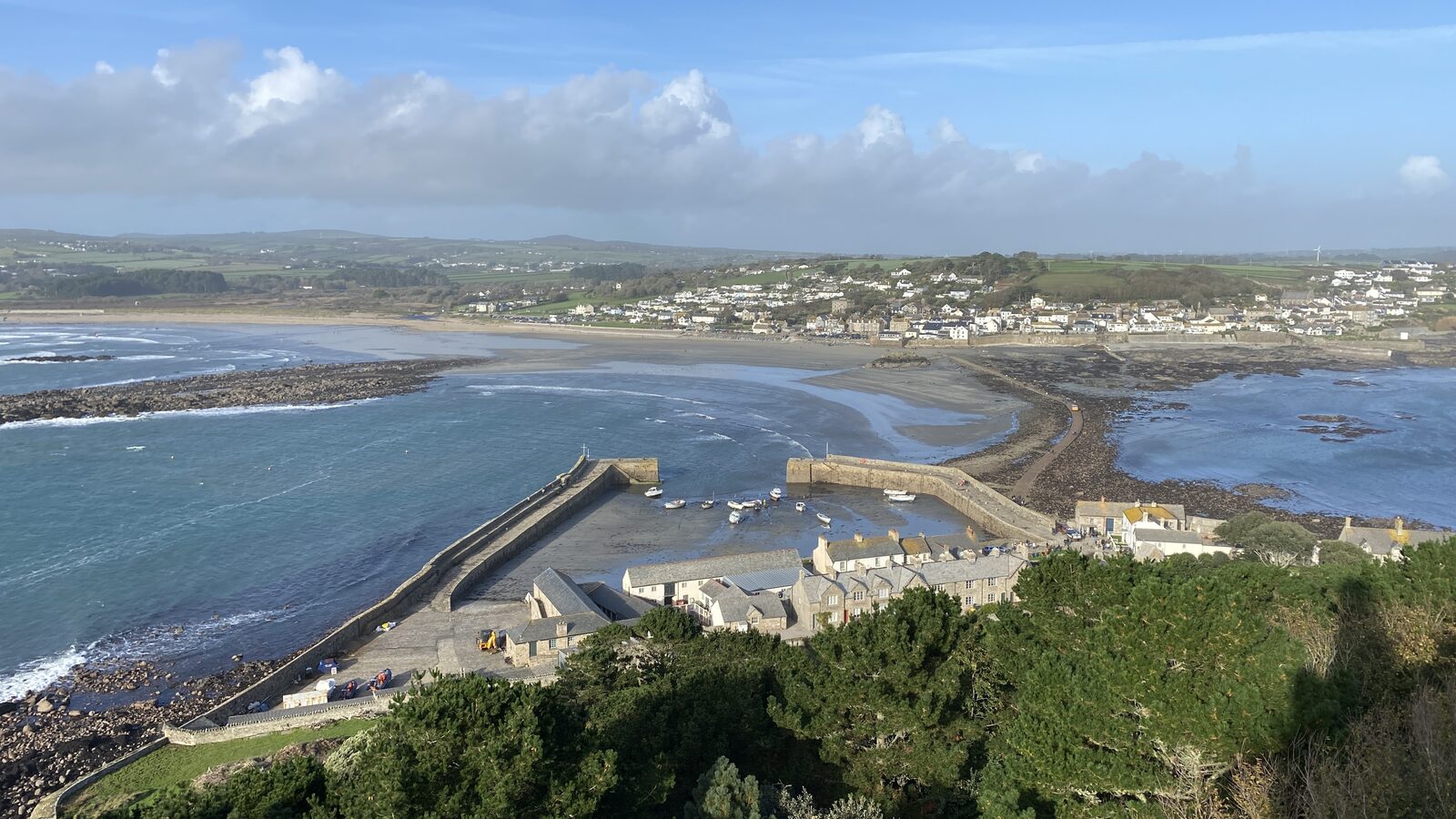 The view from St Michael's Mount looking back across the harbour and Mount's Bay to Marazion town, with the harbour walls and a few moored boats below, the long sweep of Marazion beach, and the Cornish countryside stretching inland to the horizon.