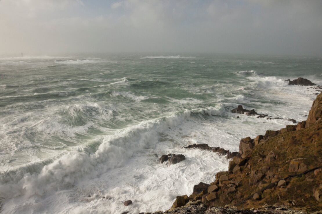 Large waves hitting the coast at Land's End in Cornwall during a storm, with the Longships Lighthouse visible offshore through the spray.