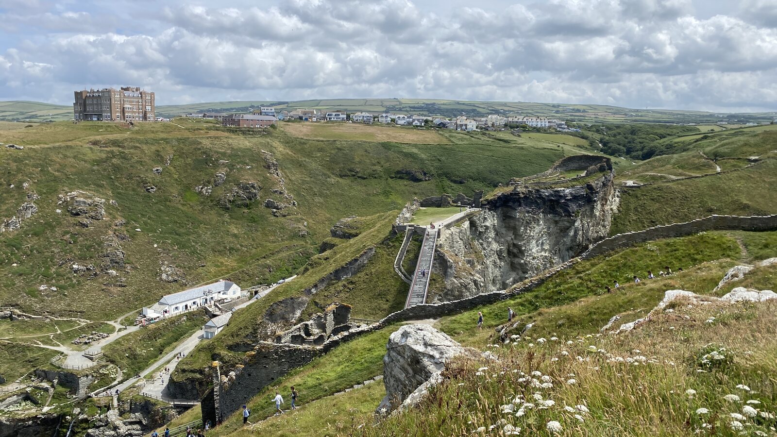 Tintagel Castle from the coast path: the ruined stone gatehouse on a rocky island connected to the mainland by a modern grey footbridge crossing a deep chasm, with a steep stepped path climbing the cliff, the visitor centre and Camelot Castle Hotel on the headland beyond, and walkers dotted across the green slopes.