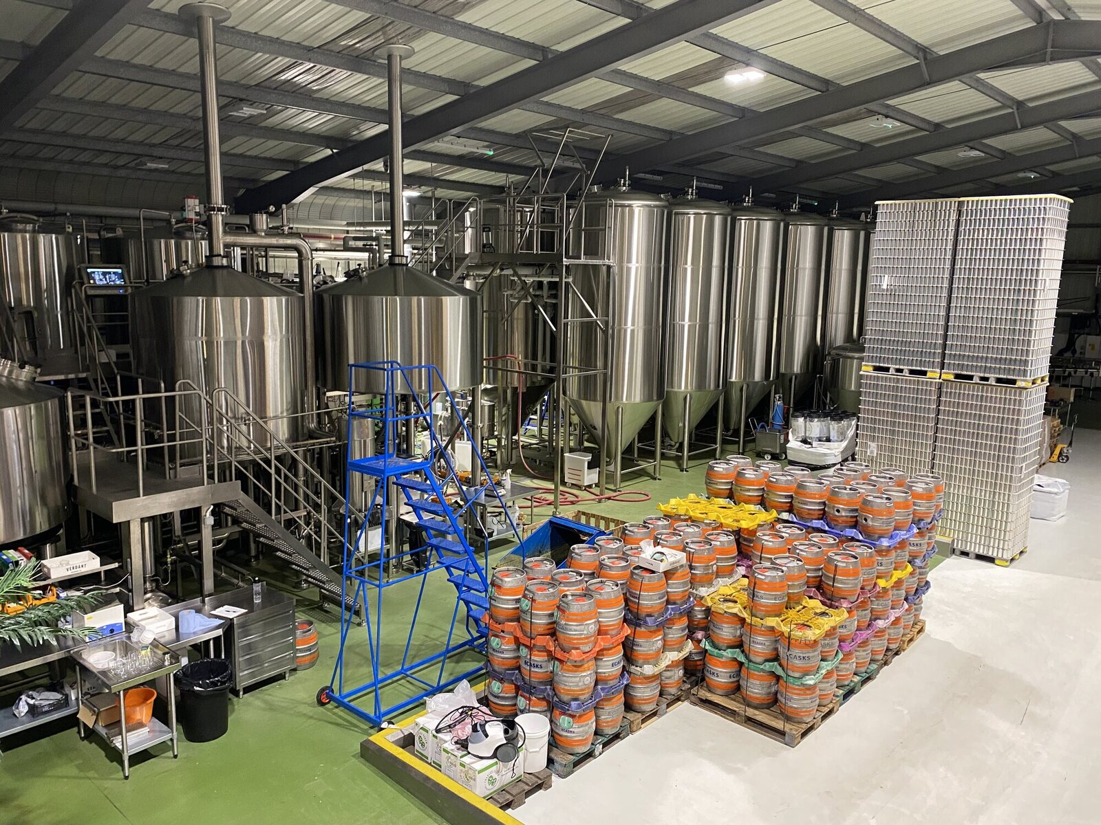 Inside Verdant Brewery in Penryn: tall stainless steel fermentation tanks rise above a green-painted brewery floor, with stacks of orange-banded firkin casks and pallets of cans in the foreground, and a blue maintenance ladder for accessing the upper tanks.