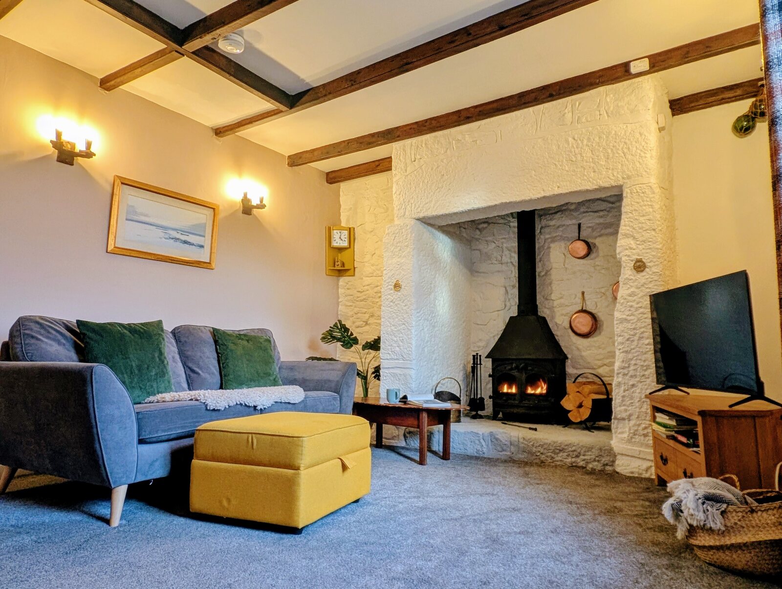 The lounge at Little Avalon, one of the Trewena cottages, with a lit wood-burner in a stone fireplace, a blue sofa with a sheepskin throw, low warm lighting from wall sconces and a yellow ottoman in front of the hearth.