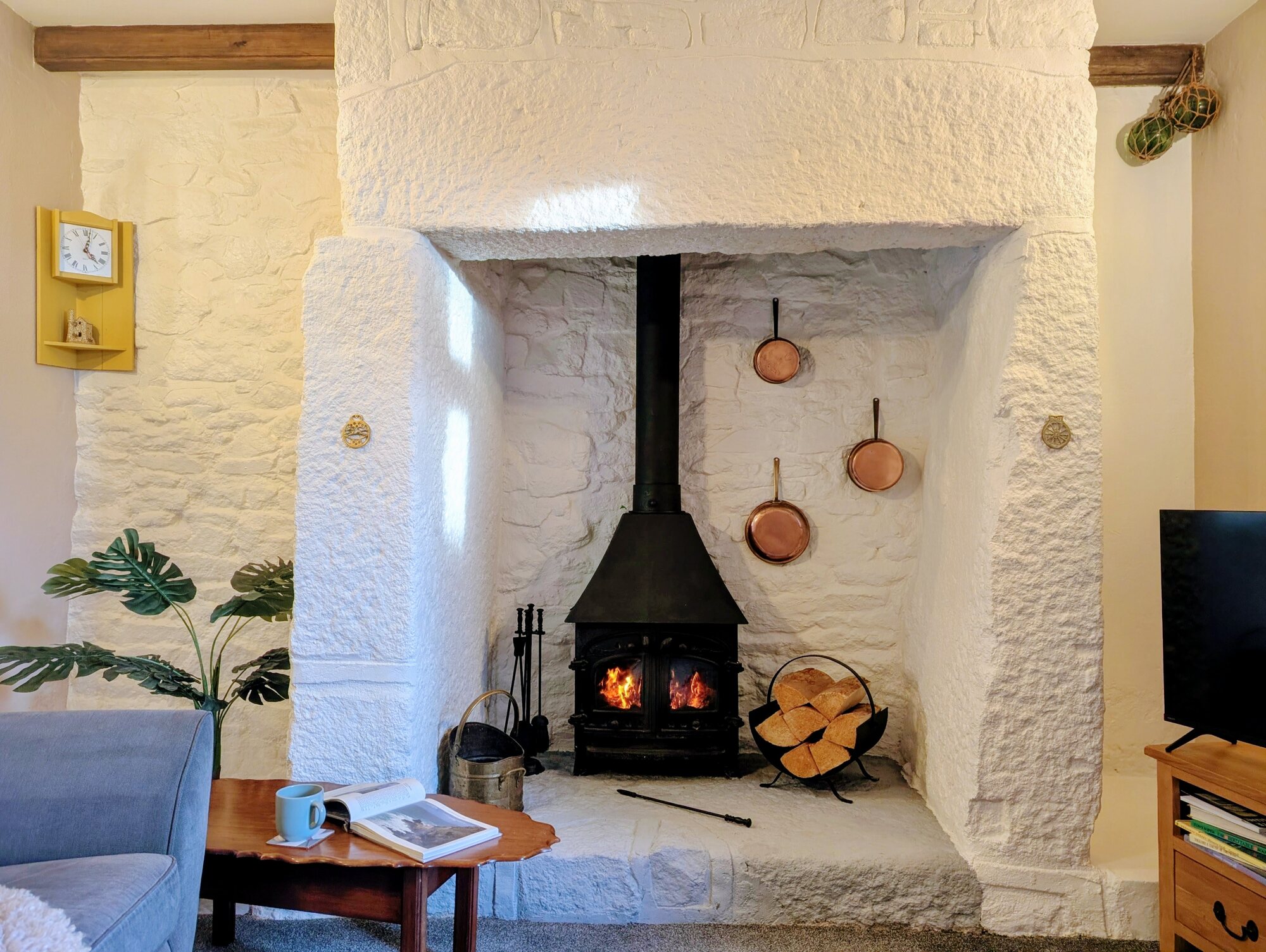 The lit wood-burner at Little Avalon, one of the Trewena cottages, set into a deep stone fireplace with copper pans on the wall, a basket of split logs, and a book and mug on the coffee table.