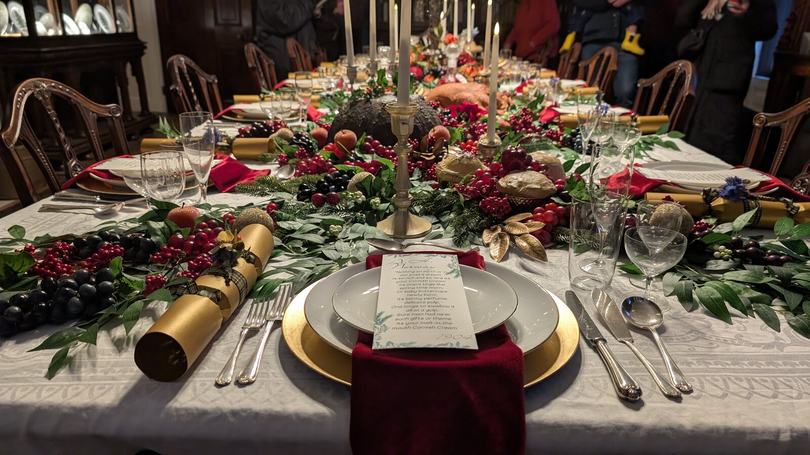 A long candlelit dining table at Trelissick House dressed for Christmas, with green and red garlands of fir, holly and berries running down the centre, gold candelabra, red velvet napkins and gilt chargers.
