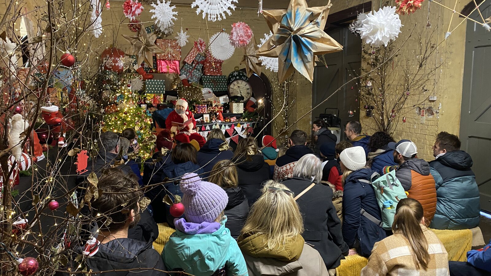 Santa in his grotto at Trelissick reading aloud to a crowd of warmly-wrapped families seated under hanging snowflakes, paper stars and Christmas lights.