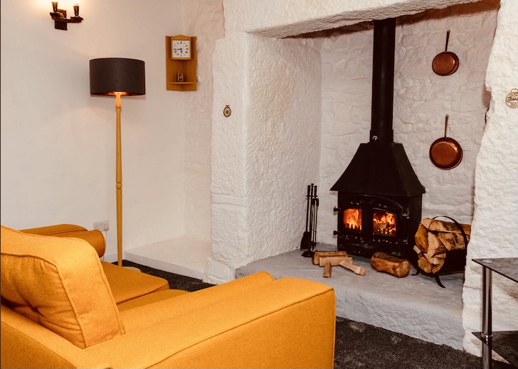A comfortable yellow sofa facing roaring fire inside a traditional Cornish cottage in winter