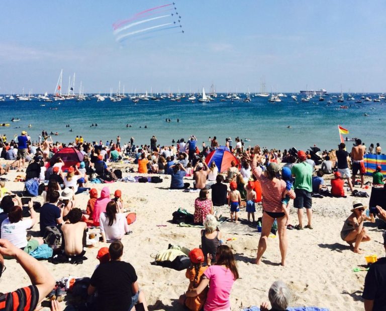 The red arrows display team fly our the coast of falmouth in cornwall watched by a packed beach of onlookers in the hot summer sun. Out to sea scores of yachts and boats are lined up to watch the aerial display.